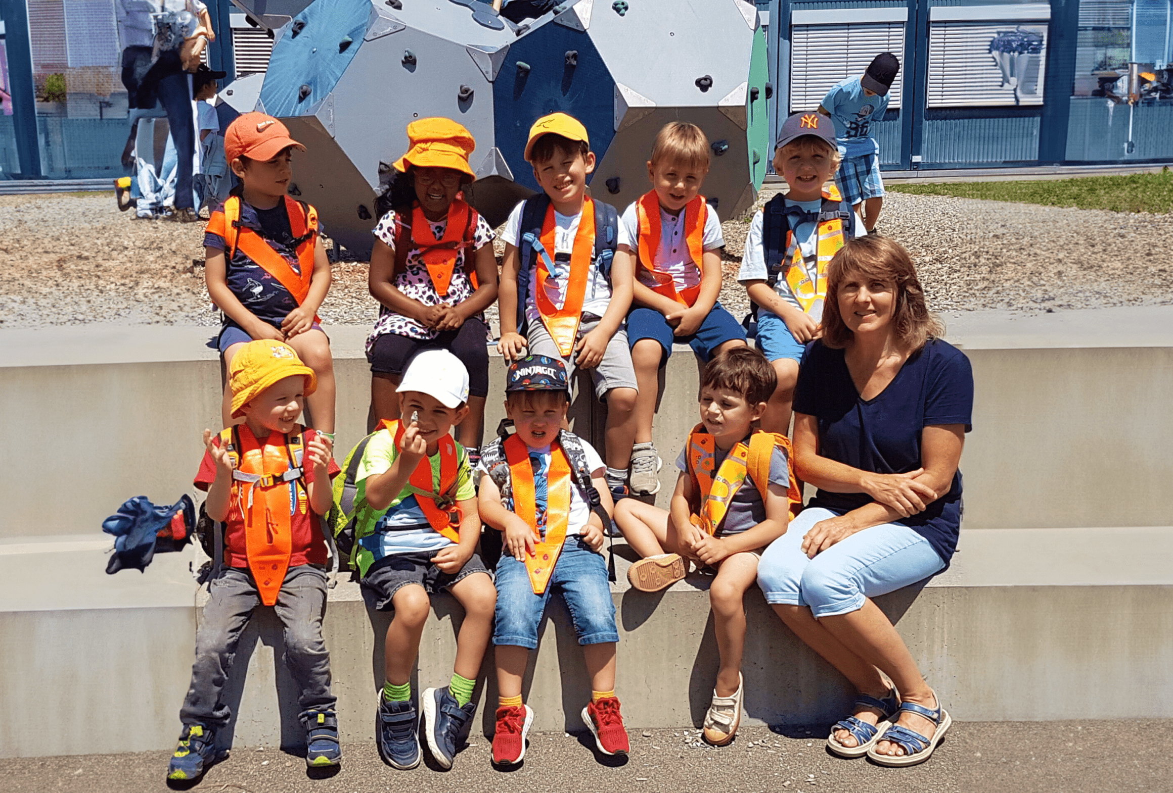 Neun Kinder mit orangen Sicherheitswesten und eine Lehrerin sitzen auf breiten Aussentreppen vor einer Kletterwand